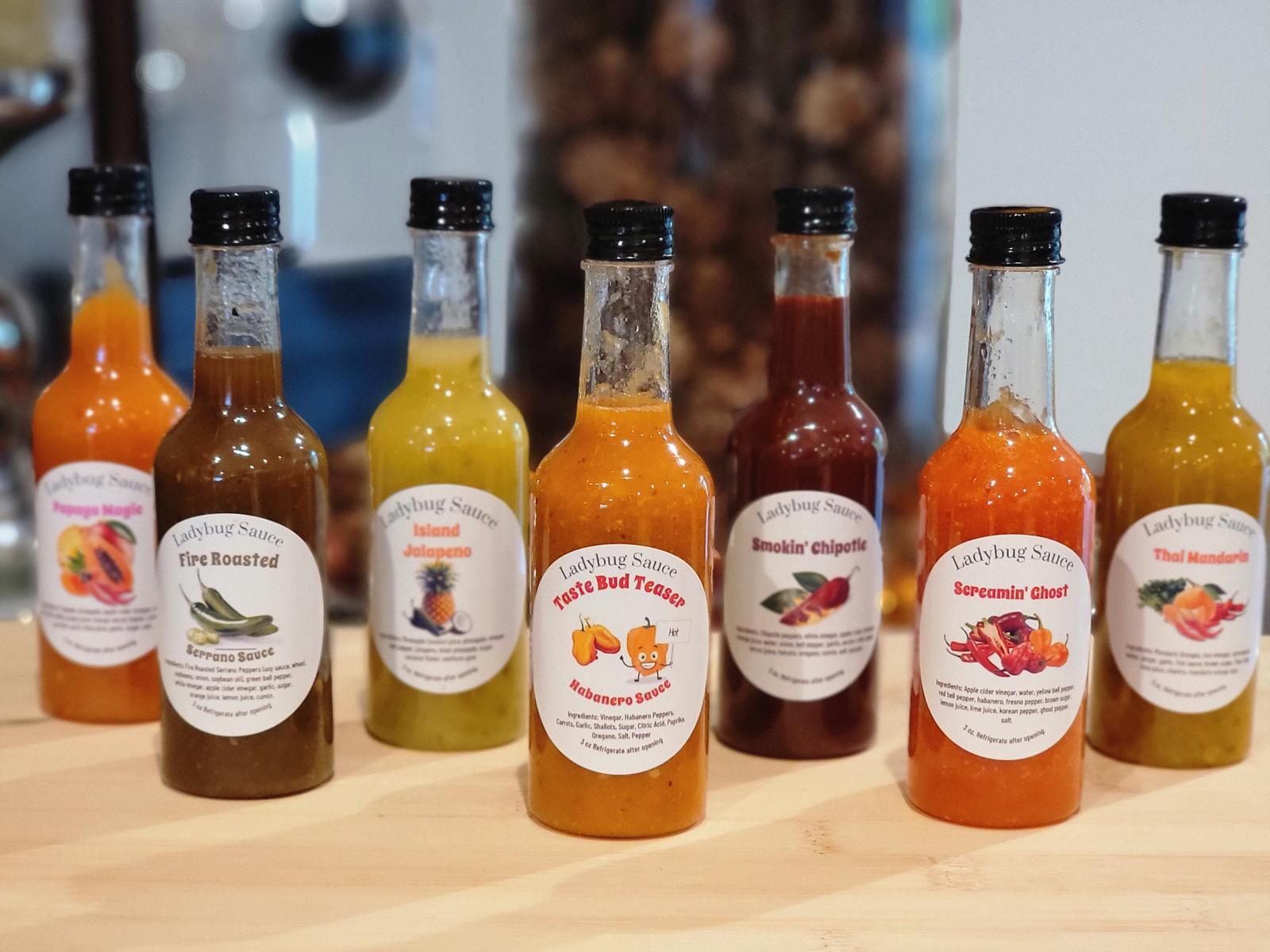 Seven Ladybug hot sauce bottles in a row: Papaya Magic, Fire Roasted, Island Jalapeno, Taste Bud Teaser, Smokin' Chipotle, Screamin' Ghost and Thai Mandarin